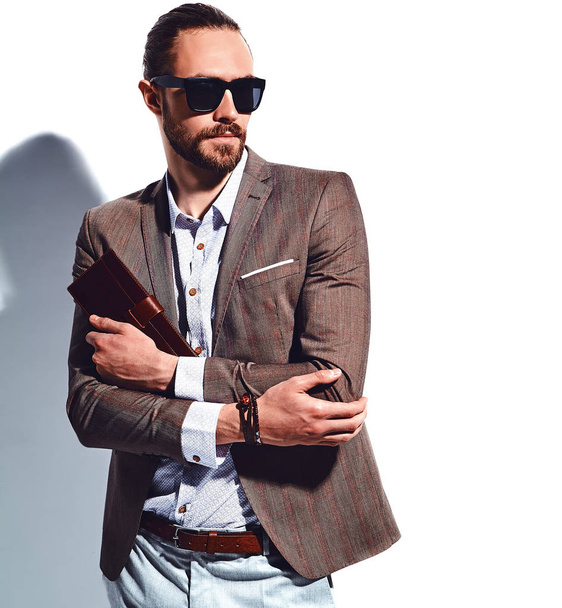 portrait of handsome fashion stylish hipster businessman model dressed in elegant brown suit in sunglasses posing near white wall in studio. Pulls out or put his leather wallet into jacket pocket - Foto, afbeelding