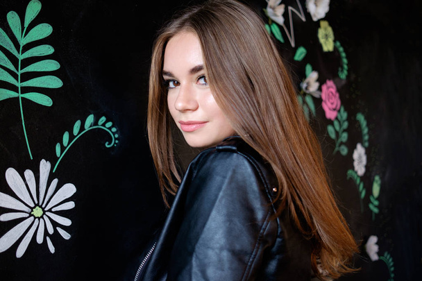 joven bonita chica morena en chaqueta de cuero sonriendo de pie cerca de la pared negra
 - Foto, Imagen