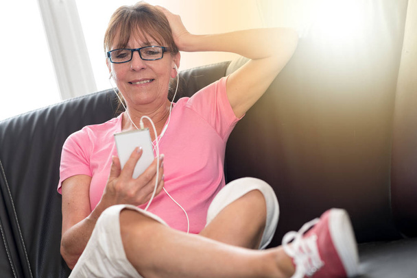 Жінка сидить на дивані і слухає музику зі своїм мобільним телефоном
 - Фото, зображення