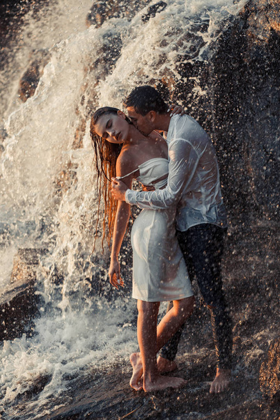Young enamored couple hugs and kisses under spray of waterfall. - Photo, Image