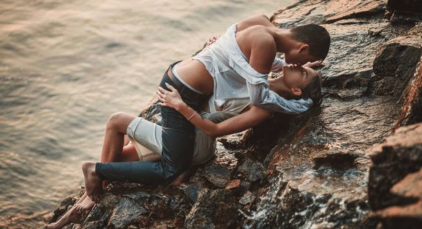Young enamored couple lies on rock, hugs and kisses under spray of waterfall.   - Zdjęcie, obraz