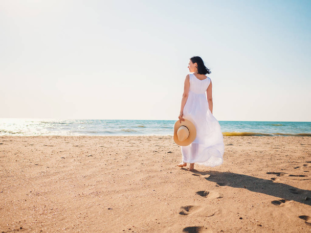 Girl wearing white maxi long dress walking barefoot on the sea shore with hat in hands. Bohemian clothing style. - Valokuva, kuva