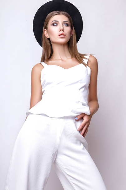 Girl in white clothes and black hat posing in studio on white background. - Zdjęcie, obraz
