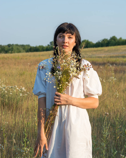 Une jeune femme dans une longue chemise brodée blanche tient un bouquet
  - Photo, image