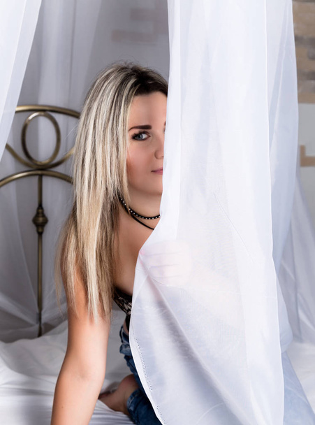 Cheerful pretty young woman sitting on a bed behind curtains and smiling - Фото, изображение