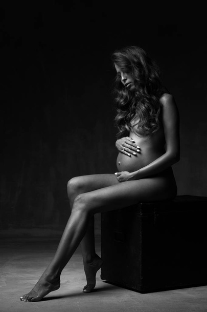 schöne junge schwangere lächelnde sexy Frau mit langen lockigen Haaren und dickem Bauch, ohne Kleidung, die im dunklen Studio auf schwarzem Würfel posiert, in schwarz und weiß - Foto, Bild