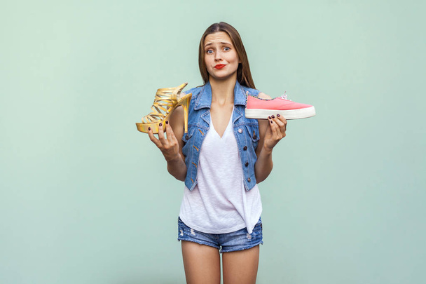 The beautiful caucasian casual girl with freckles got choosing comfortable sneakers or inconvenient but handsome high heel, thinking and looking at camera.  - Foto, Imagen
