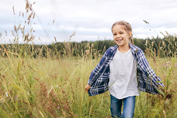 Beautiful small kid with pigtail, wearing checkered shirt and jeans, smiling pleasantly while running on green grass of meadows. Happy female child resting outdoors, having fun and good mood - Fotografie, Obrázek