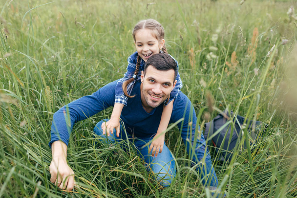 Playful attractive girl embracing her father who is sitting back to her, spending their free time together on green field. Handsome young man giving piggyback ride to his daughter in green grass - Fotó, kép