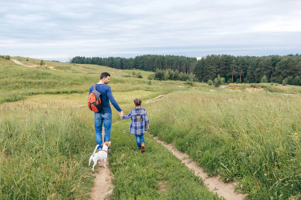 Family, parenthood, fatherhood concept. Back view of young man holding backpack and keeping hands with her little daughter and their small dog, walking on path going in forest to pick up berries - Fotografie, Obrázek