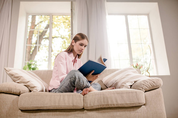 Girl at home sitting on couch reading magazine. - 写真・画像