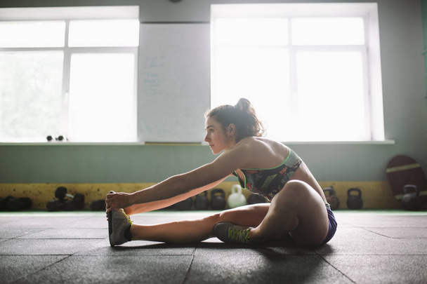 Sporty young girl doing warm-up, stretching muscles on the floor in gymnasium in backlight from window - Photo, Image