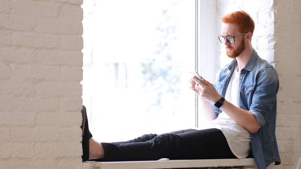 Smartphone benutzen, im Fenster sitzen, Mann mit roten Haaren und Bart - Foto, Bild