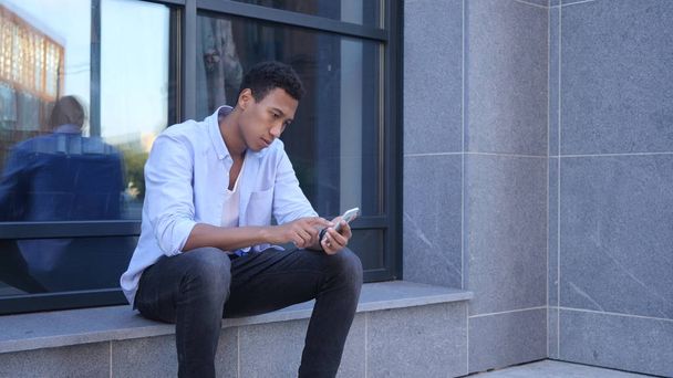 Outdoor Browsing on Smartphone, Sitting Young Black Handsome Man - Foto, Bild