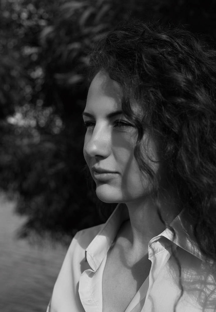 Close-up outdoor vertical black & white portrait of natural beautiful lady with long curly hair and model face dressed in shirt looking far away squinting from sunlight. Health and beauty concept. - Photo, Image