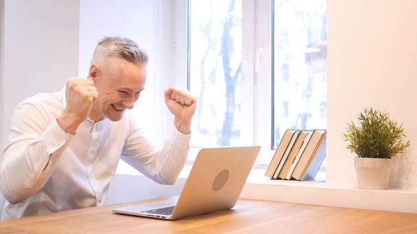Orta yaş adam kutluyor başarı dizüstü bilgisayarda çalışırken - Fotoğraf, Görsel
