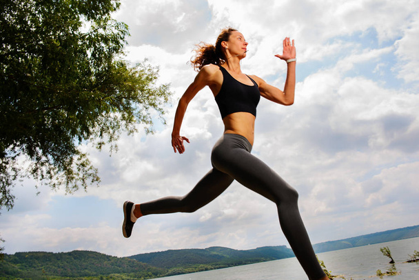 A beautiful sporty woman runing on the shore of a lake in sportswear. Girl is exercising - Photo, Image