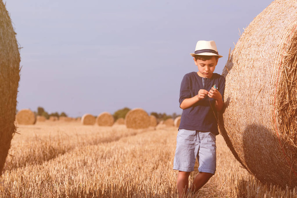 夏の夕日に干し草の山に近い帽子で若いかわいい国の少年。夏の概念、休暇。暖かい夏の日に子供とアクティブなアウトドア レジャー. - 写真・画像