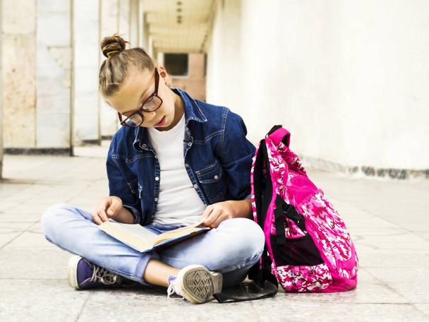 Χαριτωμένο ξανθό κορίτσι μαθήτρια διαβάζοντας ένα βιβλίο ενώ κάθεται κοντά στο σχολείο. Εκπαίδευση. - Φωτογραφία, εικόνα
