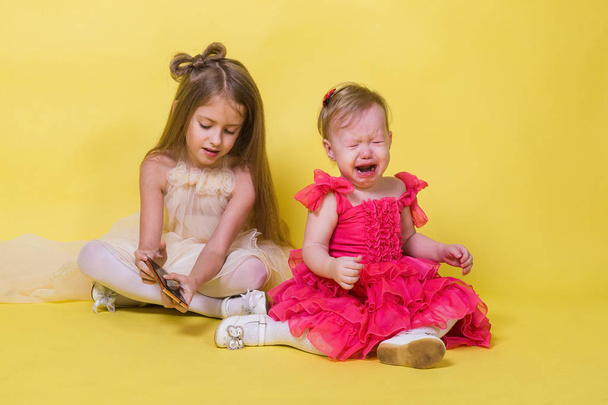 The younger sister cries, the elder keeps the phone on yellow background - Fotoğraf, Görsel