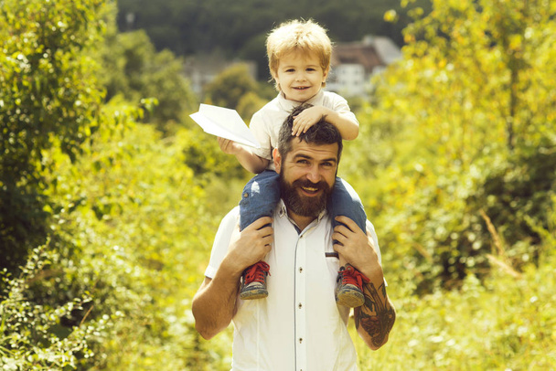 Father, son plane in nature. Bearded parent holds on shoulders cheerful cute boy with paper airplane in hands. Walking and happy childhood, hobby aviation and family travel to airport. Chidhood dream - Photo, Image