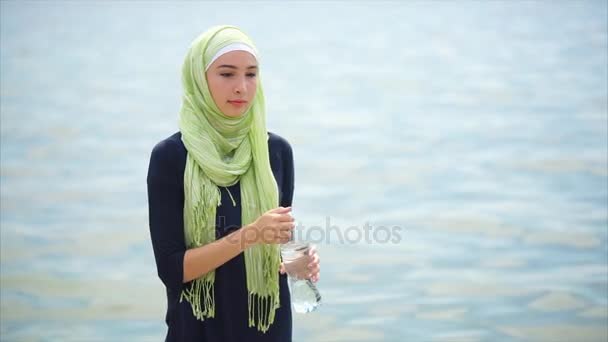 Μια νεαρή γυναίκα σε ένα hijab πίνει μεταλλικό νερό από το μπουκάλι σε μια καυτή ημέρα σε εξωτερικούς χώρους - Πλάνα, βίντεο