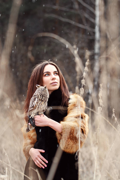 Woman in fur coat with owl on hand by first autumn snow. Beautiful brunette with long hair in nature, holding an owl. Romantic, delicate look girls - Φωτογραφία, εικόνα