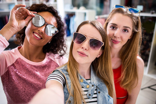 Hymyilevä tyttöystäviä yllään tyylikäs aurinkolasit hauskaa ottaa selfie matkapuhelimella, kun teet ostoksia vaatekaupassa
 - Valokuva, kuva