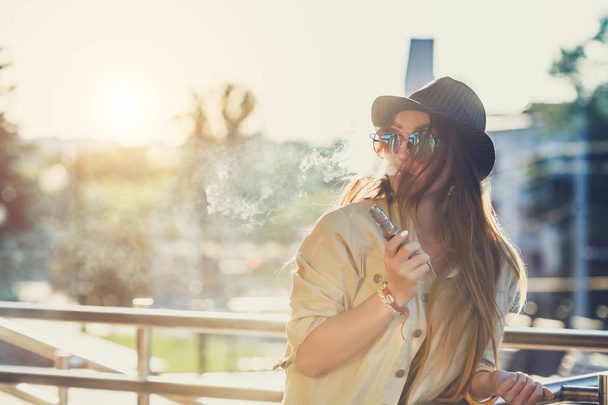 Mujer hipster bastante joven en sombrero negro vape ecig, dispositivo de vapeo al atardecer. Imagen tonificada
. - Foto, Imagen