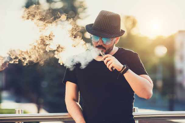 Modern happy young man in hat with a beard fun Vaporizers, In the background, the evening sunset over the city. toned image. - 写真・画像