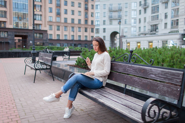 A beautiful young girl with long brown hair sitting on a bench with a book, holding eyeglasses. She left the house on a warm evening to read in the yard. The urban background. - Foto, afbeelding