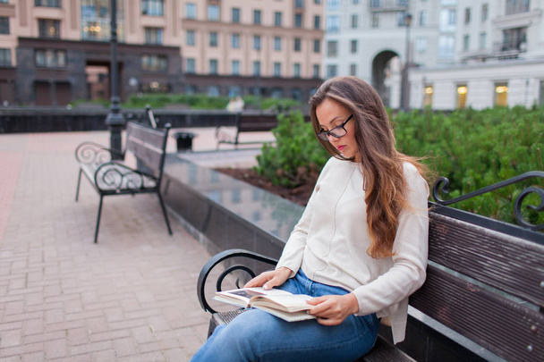Fiatal gyönyörű lány szemüveges hosszú, barna haj, ül egy padon egy könyvet. Elhagyta a házat egy meleg este, hogy olvassa el az udvaron. A városi háttér. - Fotó, kép