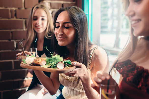 Groep meisjes met lunch in modieuze restaurant. Lachende jonge vrouw die geniet van de geur van heerlijke salade geserveerd op houten plaat - Foto, afbeelding