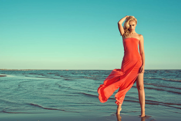 Superbe modèle blond mince sexy en robe bustier rouge avec train volant debout sur la pointe des pieds dans l'eau de mer, tenant ses cheveux ondulés soufflés par le vent
 - Photo, image