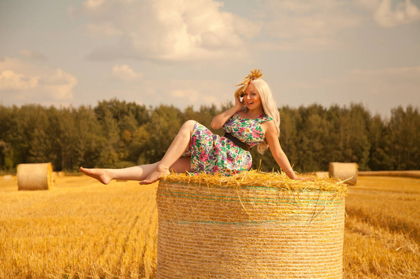 красива сільська жінка в квітковій сукні і пшенична корона стоїть біля сухого круглої соломи сіна в жовтому полі
 - Фото, зображення