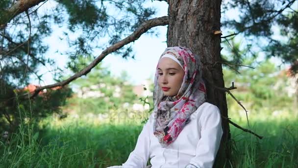 Žena v muslimský šátek sedí na trávě vedle stromu v parku - Záběry, video