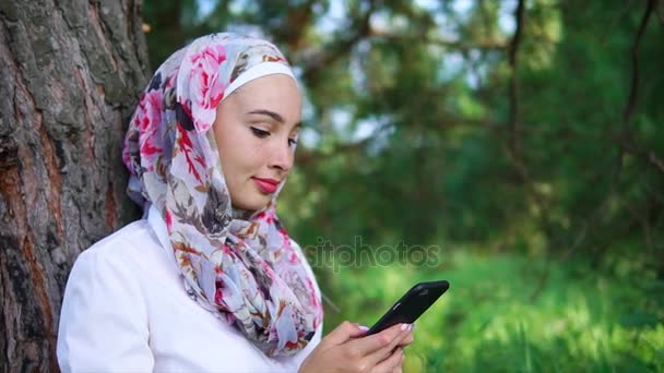 A Muslim woman with a smile is writing on the phone with a friend, in the park - Footage, Video
