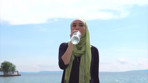 Moderne Moslimvrouw in hijab mineraal water drinkt in de buurt van de zee op straat - Video