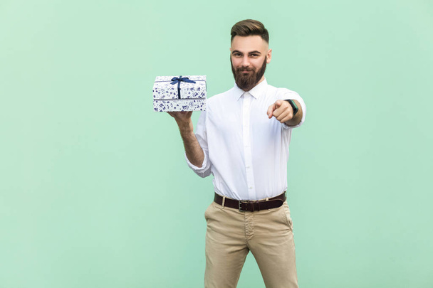 Young adult bearded man, pointing at camera and holding a gift box. Studio shot on light green background - Photo, Image