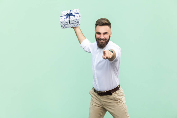 Its for you! Young adult businessman, holding gift box, pointing finger at camera and toothy smile. Isolated on light green background. Indoor, studio shot - 写真・画像