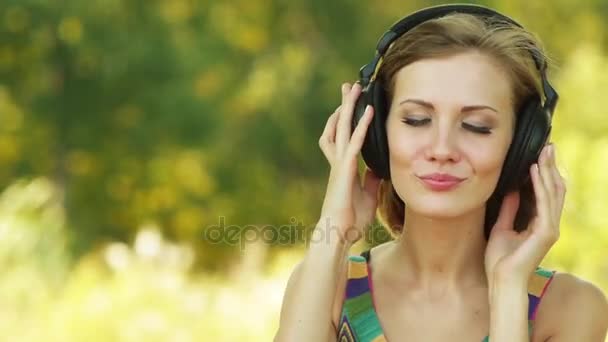 Młoda, piękna kobieta, słuchanie muzyki na słuchawkach tle zielony summer city park. - Materiał filmowy, wideo