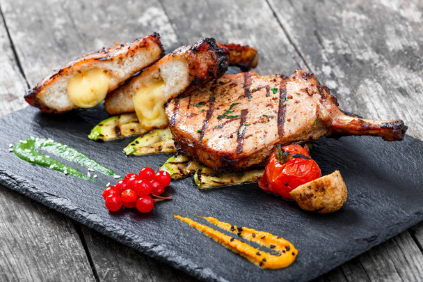 Roasted Pork steak on bone stuffed with cheese, grilled vegetables and berries on stone slate background on wooden background close up. Hot Meat Dishes. Top view with copy space - Photo, Image