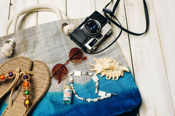  Flat lay of summer fashion with camera, slippers, sunglasses and other girl accessories on top of the bag on white wooden background - 写真・画像