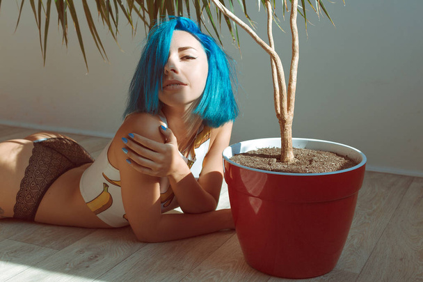 Beautiful slender girl in panties and a T-shirt with blue hair posing on the floor next to a flower in a pot - Photo, Image