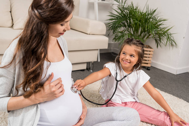 Chica escuchando vientre embarazada a través de estetoscopio
 - Foto, imagen