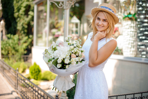 Girl in a white dress and a straw hat holds a festive bouquet - Zdjęcie, obraz
