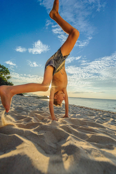 Красавчик восьмилетний мальчик на пляже выполняет акробатические эскизы
 - Фото, изображение