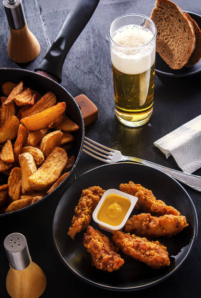 Chicken nuggets on a black plate, fried potatoes, beer and bread - Foto, Imagem