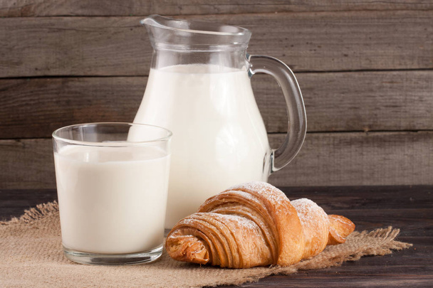 džbán a sklenice mléka s croissantem na dřevěné pozadí - Fotografie, Obrázek