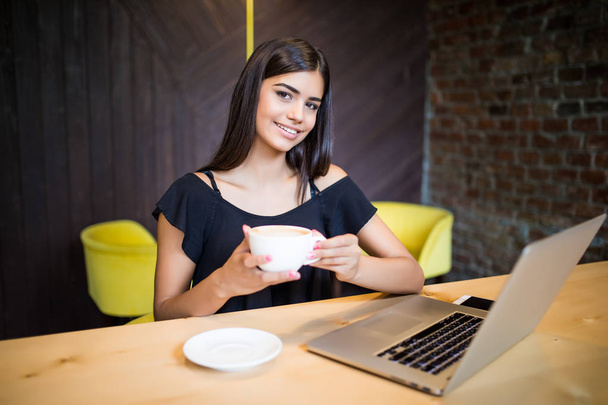 Kaunis brunette nauttia kahvia ja käyttää kannettavaa tietokonetta hymyillen aamulla
 - Valokuva, kuva
